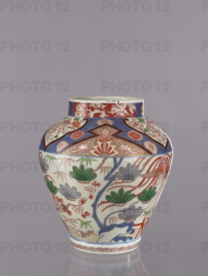 Vase tsubo, Japanese, decorated with flowers, plant ornaments, phoenixes, geometric ornaments, colored porcelain, Imari, pine trees; ceramics, Arita, Japan; ca. 1720–1740; height 43 cm; vessel., A rounded ceramic vase with a short neck sits against a plain gray background, decorated with intricate painted patterns in red, blue, green, and gray. The upper band features geometric and floral motifs in red and blue with cream highlights, while the main body shows stylized plants and flowers with green leaves, gray petals, and red branches on a cream-colored ground. A blue winding trunk or branch motif runs vertically among the floral designs, and a narrow decorative border encircles the base in red and cream. The surface appears glossy and smooth, and the overall color palette includes red, blue, green, gray, cream, and touches of brown.