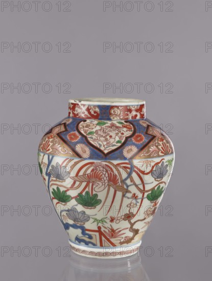Vase tsubo, Japanese, ca. 1720–1740, Arita, Japan. Colored porcelain Imari, ceramics; height 43 cm. Decorated with flowers, plant ornaments, pine trees, phoenixes, and geometric ornaments., A rounded ceramic vase with a short neck displayed against a plain muted gray background; the vase has a glossy white base color decorated with intricate painted motifs in red, blue, green, and gold tones, including floral shapes, stylized leaves, and flowing ribbon-like lines; a band of deep blue and red panels with ornamental patterns encircles the upper body beneath the neck, while the central field shows a large stylized floral element with long curved petals in red and gold surrounded by green leaf clusters and blue-gray flower forms; the lower rim features a repeating circular spiral pattern in red and the neck rim is edged with red and blue ornamental designs.