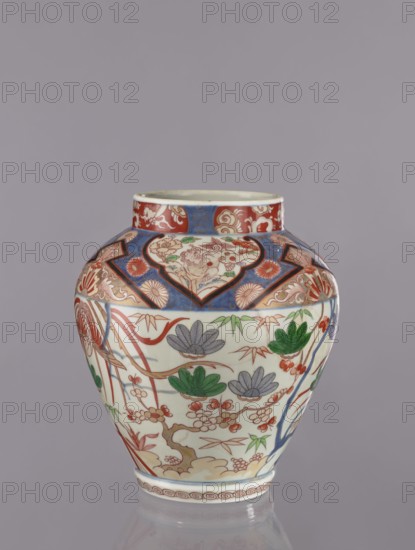 Vase tsubo, Japanese, Imari colored porcelain, Arita, ca. 1720–1740, vessel with phoenixes, flowers, pine trees, geometric and plant ornaments, height 28.5 cm, The image shows a ceramic vase with a rounded body and short neck against a plain gray background. The vase is decorated with ornate floral and botanical motifs in colors including red, blue, green, and gray on a white base. The upper section features patterned panels with red and blue backgrounds and decorative swirls, while the main body displays stylized flowers, leaves, and branches in shades of red, green, and gray with fine linear detailing. A band of repeating circular motifs encircles the base, and the rim has a narrow decorated border. The overall surface combines bold colored areas with delicate line work and a glossy finish.