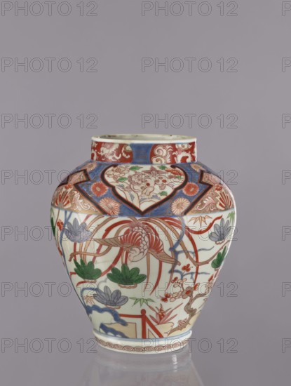 Vase tsubo, Japanese, ca. 1720–1740, Arita, Japan. Colored porcelain Imari, decorated with phoenixes, flowers, pine trees, plant and geometric ornaments. Vessel, height 28.5 cm., A rounded porcelain vase with a short neck sits against a plain gray background, decorated in a palette of red, blue, green, and cream with touches of brown and purple. The upper neck band features a red and blue patterned border with floral and scroll motifs, while the main body displays a large central bird with flowing red feathers surrounded by stylized flowers and leaves in green, purple, and blue. Heart-shaped cartouches and geometric panels in blue and red frame floral sprays and cloud-like forms, and the lower rim has a narrow decorative band in red. The glaze is glossy and the vase shows fine painted detailing throughout.