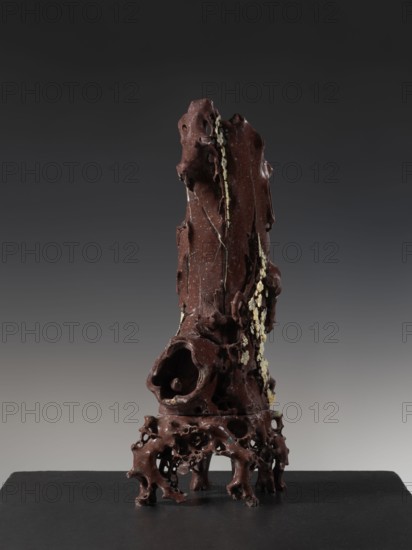 Vase on base, Chinese art, Chinese culture, Qing 1644–1911, huaniao style, plum trees, birds and flowers, brown soapstone, late 19th century, China, vessel, height 43.0 cm, A tall, irregular vertical sculpture with an organic, weathered surface stands on a dark flat base; the sculpture is primarily deep brown with areas of lighter brown and small pale yellowish beige clusters along one side, and it has many hollowed cavities, rough textured ridges, and drip-like formations; the lower portion flares into a porous, root-like support with rounded openings and uneven legs, while the upper portion tapers and features knobby protrusions and narrow vertical grooves; the background is a smooth gradient from dark gray at the top to lighter gray toward the bottom.