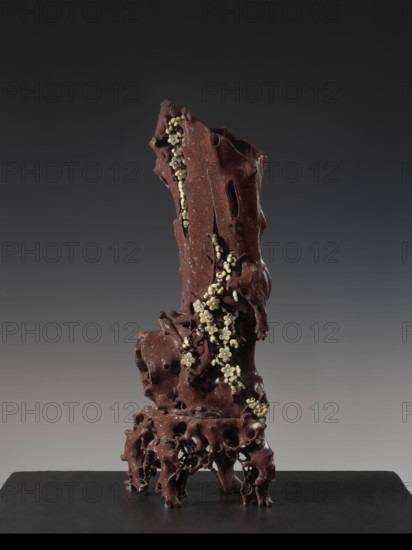 Vase on base, Chinese art, Chinese culture, Qing 1644–1911, huaniao style, plum trees, birds and flowers, brown soapstone, late 19th century, China, vessel, height 43.0 cm, A tall sculptural object with an irregular, porous form stands on a flat dark surface against a muted gray background; the sculpture is predominantly deep reddish-brown with a rough, pitted texture and flowing, carved cavities, and a winding cluster of small pale cream-colored blossoms and rounded buds ascends along its front, contrasting with the darker material.