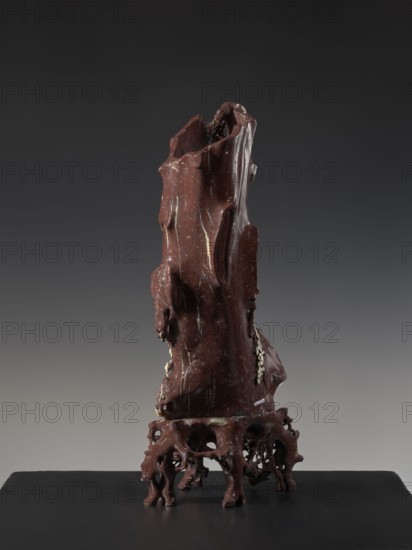 Vase on base, Chinese, Qing 1644–1911, huaniao style; decorated with plum trees, birds and flowers; brown soapstone; late 19th century, China; vessel; height 43.0 cm, A tall, irregularly shaped object with a glossy, deep reddish-brown surface standing on an openwork base of similar color; the object has a rough, carved appearance with smooth, flowing contours and several cavities and protrusions, with visible pale streaks and small pale flecks running across its surface; the base is organic and root-like with multiple arching supports and hollowed openings; the background is a smooth gradient ranging from dark charcoal at the top to a lighter gray toward the bottom, and the platform beneath the object is a flat black surface.