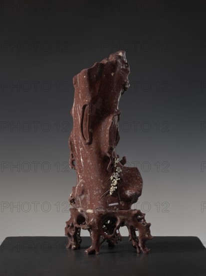 Vase on base, Chinese, Qing 1644–1911, huaniao style, decorated with plum trees, birds and flowers, brown soapstone, late 19th century, China; vessel, height 43.0 cm, A tall, irregularly shaped carved object in deep reddish-brown with lighter speckles stands on a dark surface; its form is organic with smooth and rough contours, several carved recesses and protrusions, and a cluster of small pale elements attached to one side. The base is intricate and root-like with multiple openings and rounded legs in the same reddish-brown color. The background is a smooth gradient of dark gray to lighter gray. The visible colors are reddish-brown, pale off-white, dark gray, and black.
