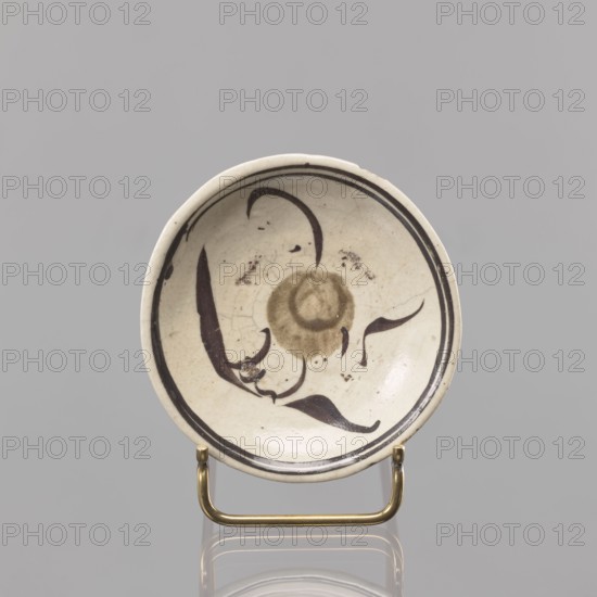Plate, Southeast Asia culture, floral decorations, ceramics, trace of a Chinese inscription on the bottom, 18th–19th century, Southeast Asia place of origin, vessel, height 2 cm, diameter 10.5 cm, A shallow round ceramic dish with a cream-colored surface and a glossy finish sits on a small brass stand; the interior shows a decorative motif in dark brown and muted olive tones forming a central circular shape and flowing, curved brushstrokes that suggest organic, leaf-like forms, with a dark brown double border near the rim; the glaze shows fine crackling and a few small dark spots, and the background surrounding the dish is a uniform light gray.
