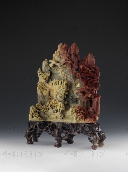 Mountain Landscape with Architecture on a Base - Bas-Relief, Chinese, Qing 1644–1911, Mid-19th century. Sculpture; materials/technique: bas-relief. Subject: mountain landscape with architecture; decoration: landscape motifs, architectural elements. Dimensions: height 33.5 cm, width 27.5 cm., A carved stone sculpture resting on an intricately carved dark wooden stand, showing a vertical rocky landscape with layered, porous textures and delicate pierced detailing; the stone transitions in color from pale yellow and beige on the left through gray tones in the center to deep red and rust on the right, with carved overhangs and ledges resembling terraces and clustered organic forms, and a small pavilion-like carved structure nestled among the red area, all set against a dark gray gradient background.