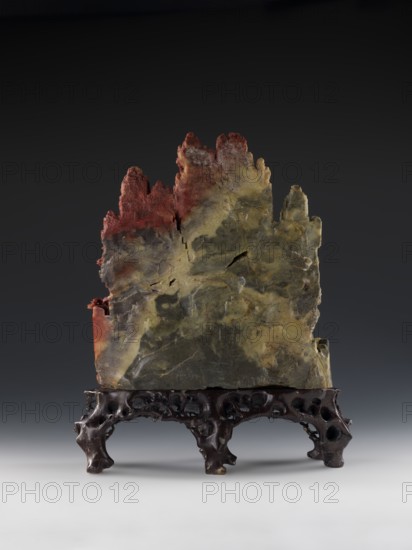 Mountain Landscape with Architecture on a Base - Bas-relief. Chinese; Qing dynasty 1644–1911. Mid-19th century. Sculpture in stone, depicting mountain landscape with architecture; decorative landscape motifs. Height 33.5 cm; width 27.5 cm., A vertically oriented jagged stone slab with an irregular top edge sits on an ornate dark wooden stand; the stone shows a mix of colors including greenish-gray, olive, yellowish-beige, and areas of reddish-brown toward the top and left side, with visible cracks, rough textures, and mottled patterns across its surface, while the stand has a glossy dark brown finish with carved, organic openwork shapes and curved legs against a neutral gray to black gradient background.