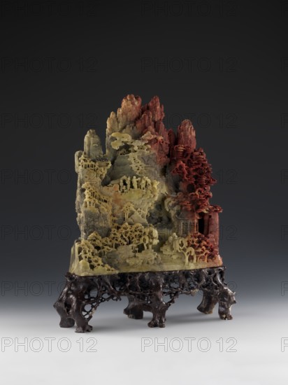 Mountain Landscape with Architecture on a Base - Bas-Relief. Unknown creator craftsman. Chinese, Qing dynasty 1644–1911, mid-19th century. Sculpture, height 33.5 cm, width 27.5 cm. Themes: decorations, architecture, mountain landscapes, landscape motifs., A carved stone sculpture resembling a mountainous landscape sits on an intricately carved dark wooden stand. The sculpture shows layered rocky peaks and ledges with detailed miniature trees, pavilions, and terraces carved into the stone. The left and central areas of the stone are pale yellow and gray with delicate openwork foliage and rounded tree forms, while the right side transitions to reddish tones with denser carved trees and architectural details. The wooden base is dark brown with a porous, root-like texture and multiple short legs, and the background is a gradient from dark gray at the top to lighter gray near the bottom.