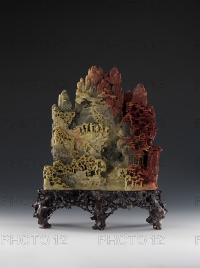 Mountain landscape with architecture on a base — bas-relief., craftsman. Chinese, Qing dynasty 1644–1911, mid-19th century. Decoration depicting mountain landscapes and architectural motifs. Sculpture; height 33.5 cm, width 27.5 cm., A carved, multi-tiered landscape sculpture sits on an ornate dark brown stand, with a range of earthy colors including pale yellow, grey, greenish tones, and deep red visible across the stone. The sculpture rises in jagged peaks and terraces, with intricately carved tree forms, layered rock ledges, and small pavilion-like structures integrated into the slopes. Delicate, lacy foliage and rounded canopy shapes appear across the terraces, and there are tiny carved figures and groupings of human-like silhouettes placed on ledges and pathways. The right side of the carving transitions to richer red hues and features more clustered, layered tree canopies, while the left and central areas are predominantly pale yellow and grey with textured rock faces and recesse...