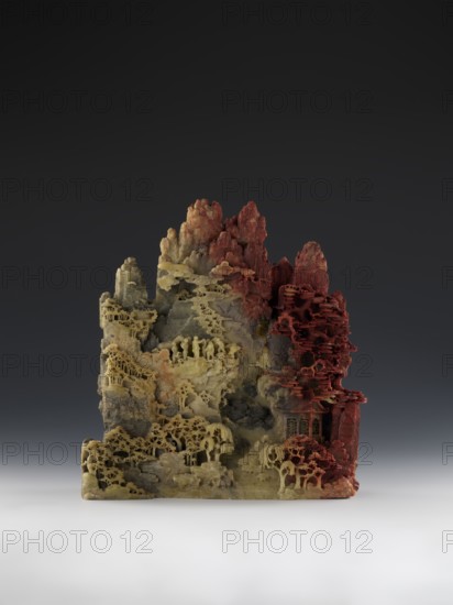 Mountain landscape with architecture on a base — bas-relief; creator, mid-19th century Qing dynasty, 1644–1911; Chinese; sculpture; decorations; architecture; mountain landscapes; landscape motifs; height 33.5 cm, width 27.5 cm., A carved landscape-like object with textured rock formations and layered terraces in shades of yellow, beige, gray, and red, featuring intricate cutaway patterns that form organic, tree-like shapes and small ledges; the piece rises from a rectangular base and shows clusters of rounded and jagged peaks, with pale yellow and beige areas concentrated on the left and lower portions and deep red tones on the right and upper peaks, set against a dark gradient background that transitions to a lighter surface under the base.