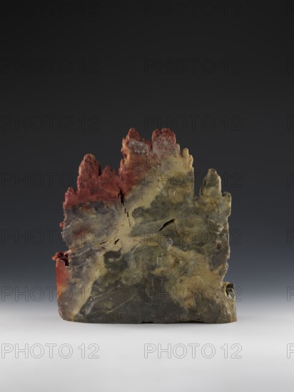 Mountain Landscape with Architecture on a Base - Bas-Relief, Chinese, Qing 1644–1911, mid-19th century. Sculpture depicting mountain landscapes with architectural decorations; height 33.5 cm, width 27.5 cm., A rough, irregular vertical stone slab with jagged peaks and a weathered surface sits on a smooth light-gray base against a dark-gray to nearly black gradient background; the stone shows a mix of colors including muted green, olive, yellowish-beige, dark gray, and areas of rusty red concentrated near the top and along one side, with visible cracks, pitted textures, and layered striations giving it a rugged, natural appearance.