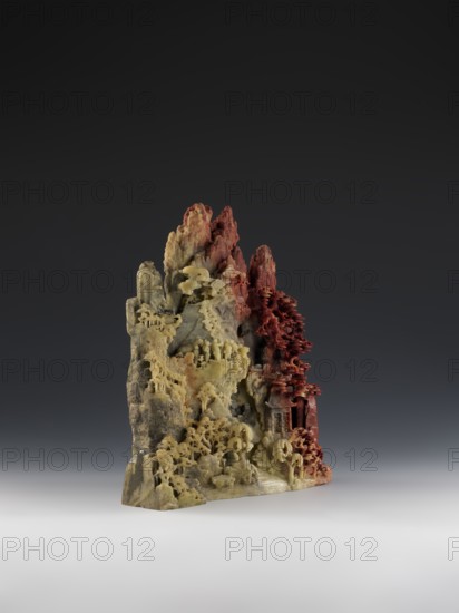 Mountain landscape with architecture on a base — bas-relief; maker:, role: craftsman; Chinese; Qing dynasty 1644–1911; mid-19th century; subject: mountain landscapes with architectural decoration; material: sculpture; height 33.5 cm; width 27.5 cm., A carved stone sculpture resembling a mountainous landscape with detailed textures and layered forms, showing a transition of colors from pale yellow and beige at the base and left side to reddish hues on the peaks and right side, with intricate carved shapes that suggest terraces, overhangs, and clustered formations, set against a dark gray to black gradient background and resting on a smooth light gray surface.