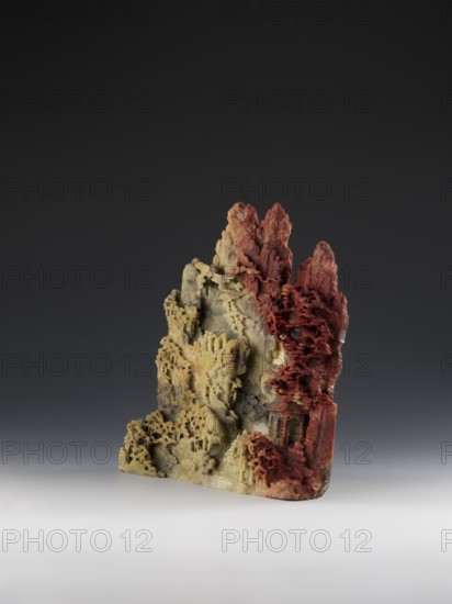 Mountain landscape with architecture on a base — bas-relief. Unknown craftsman. Chinese, Qing dynasty 1644–1911. Mid-19th century. Carved decoration showing architecture and mountain landscape motifs. Sculpture; height 33.5 cm, width 27.5 cm., A carved vertical stone-like sculpture with an irregular, jagged silhouette sits against a dark gray to black gradient background and a light gray surface; the piece shows a transition of colors from muted yellowish-green on one side to deep reddish tones on the other, with areas of pale cream and subtle gray mixed in. The surface is intricately textured with layered, porous formations resembling rugged rock faces and delicate, lacy patterns, and there are carved recesses and protrusions that create shadowed hollows and fine ridges. The reddish section has denser, more clustered, almost tree-like or organic filigree details compared with the more open, eroded appearance of the yellow-green side, and small terrace-like ledges and cavernous pockets...