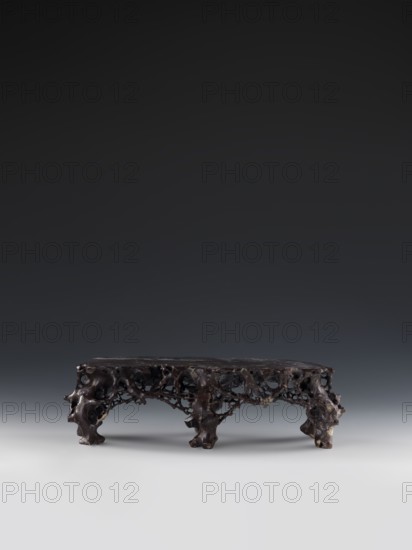 Mountain Landscape with Architecture on a Base - Bas-Relief, Chinese, Qing 1644–1911, mid-19th century. Subject: mountain landscape with architecture; style: decorations, landscape motifs, architecture. Medium: sculpture bas-relief. Dimensions: height 33.5 cm, width 27.5 cm., A low, irregularly shaped wooden stand with an organic, porous structure sits centered on a smooth surface; the stand is dark brown to nearly black with variations of lighter brown and some pale tan spots, and it has a flat top supported by a group of uneven, knobbly legs and a lattice of natural holes and tunnels. The background is a smooth gradient transitioning from dark gray at the top to a lighter gray near the middle and pale gray at the bottom, and soft shadows fall beneath the stand.