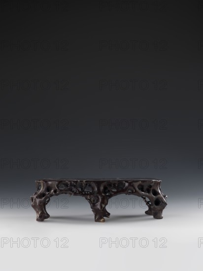 Mountain Landscape with Architecture on a Base — Bas-relief; Unknown craftsman; Mid-19th century Qing dynasty, 1644–1911; Chinese; Sculpture; Decorations; Architecture; Mountain landscapes; Landscape motifs; Height 33.5 cm; Width 27.5 cm, A low carved wooden stand with an irregular, organic form, showing a dark brown surface with areas of lighter brown where the finish is thin; the stand has a rectangular top supported by a group of sculpted, porous legs and arches that create multiple negative spaces, and the overall texture appears smooth in places and pitted in others; the background gradates from dark gray at the top to lighter gray near the base, and the surface beneath the stand is pale gray.
