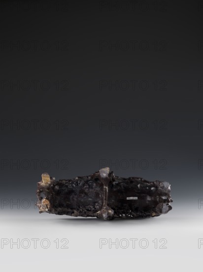 Mountain landscape with architecture on a base; bas-relief., craftsman. Chinese; Qing dynasty 1644–1911. Decorations, architecture, mountain landscapes, landscape motifs. Mid-19th century. Sculpture. Height 33.5 cm; width 27.5 cm., A dark irregularly shaped object with a rough, pitted surface sits centered against a gradient background that transitions from dark gray at the top to light gray at the bottom; the object appears primarily dark brown to nearly black with areas of lighter brown and amber tones on some protrusions, and the scene includes subtle highlights and shadows on the object's textured surface.