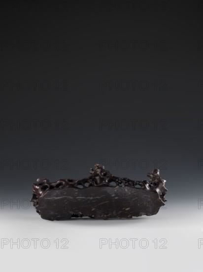 Mountain landscape with architecture on a base; bas-relief., craftsman. Medium: sculpture. Chinese, Qing dynasty 1644–1911. Decorations: architecture, mountain landscapes, landscape motifs. Date: mid-19th century. Dimensions: height 33.5 cm, width 27.5 cm., A dark brown, irregularly shaped object with a rough, textured surface sits centered on a neutral light gray surface against a gradient dark gray background; the object has a horizontal, elongated form with porous, hole-filled upper edges and knobby protrusions at either end, and its surface shows small lighter brown and beige flecks and subtle polished areas that reflect light.