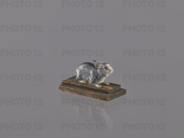 Rabbit, ceramic figurine/sculpture of an animal rabbit, ceramicist, Japanese, Meiji period 1868–1912, early 20th century, height 2.2 cm, width 4.1 cm; inscriptions/repossessions/, A small sculpted animal figure in shades of gray and white sits on a group of wooden blocks in brown tones; the figure has a glossy surface and a subtle reflection on the smooth gray background, with visible contours of ears, face, and paws, and the wooden base shows darker and lighter brown grain and worn edges.