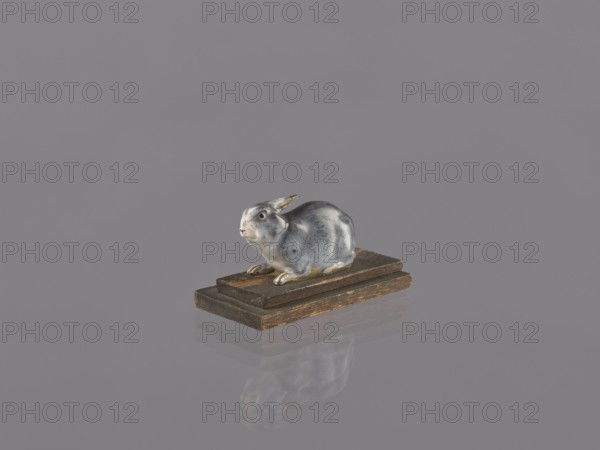 Rabbit, ceramic figurine/sculpture of a rabbit, unknown ceramicist, Japan, Meiji period 1868–1912, early 20th century; height 2.2 cm, width 4.1 cm; subject: animals/rabbits; Japanese art/culture., A small ceramic rabbit figurine with shades of gray, white, and a touch of brown sits on a pair of stacked wooden blocks that are brown with darker worn edges, all set against a smooth gray background with a faint reflection of the rabbit and blocks visible below.