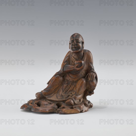 Sage - monk, Chinese art, Chinese culture, Qing 1644–1911, figurine of a sage/monk, 19th century, China, sculpture, height 8.4 cm, A carved wooden figure of a seated person with rounded facial features and a smiling expression, wearing flowing robes, posed with one arm bent holding a small rounded object and the other arm resting on a rounded support; the figure sits on an irregular carved base that resembles natural rock. The visible colors are brown tones in the wood with darker brown areas from patina and some lighter brown highlights, and a neutral gray to white gradient background.