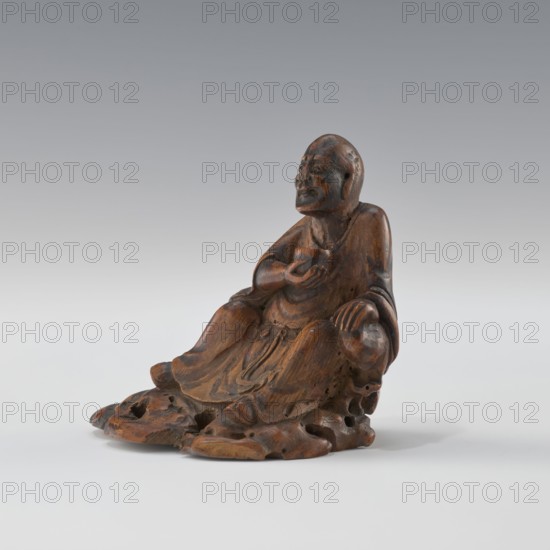 Sage - monk, unknown sculptor, Chinese art, Chinese culture, Qing 1644–1911, figurine of a sage/monk, 19th century China, sculpture, height 8.4 cm, A small carved wooden figure sits on an irregular base, showing a seated person with folded legs and draped robes. The figure's surface is brown with darker brown grain and some lighter brown highlights, and there are visible carved folds and rounded contours in the clothing. The person has a rounded head with carved facial features, an arm bent and holding a small rounded object near the chest, and the other hand resting on the knee. The base and lower robes show carved openings and textured, organic shapes, and the background is a smooth gradient of gray to off-white.