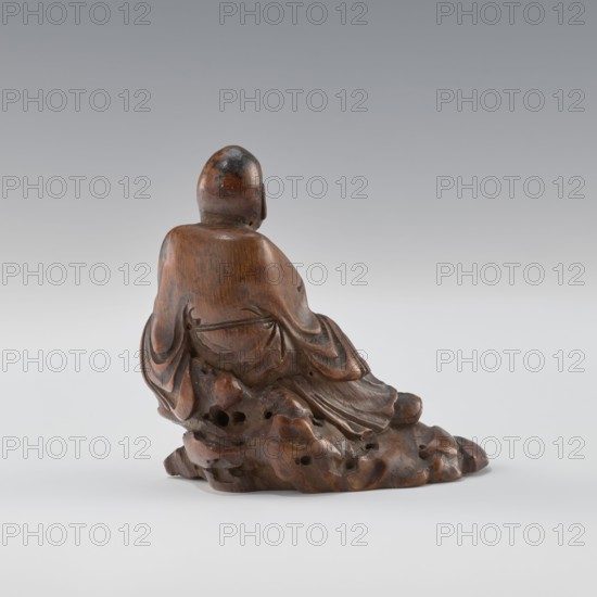 Sage — monk, unknown sculptor; Chinese; Qing dynasty 1644–1911; 19th century; porcelain/figurine sculpture depicting a sage/monk; height 8.4 cm., A carved wooden figure viewed from behind sitting on an irregular rocky base, the figure wearing flowing drapery with visible carved folds and rounded shoulders, a smooth rounded head with darker patches, and the wood showing warm brown and darker brown tones with subtle highlights against a pale gray to white gradient background.
