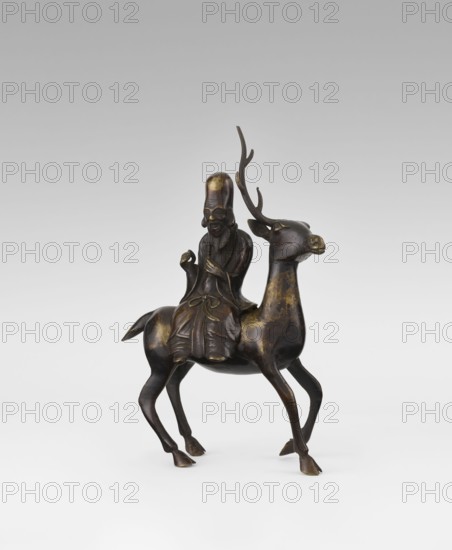Shoulao on a deer — incense burner, sculptor unknown, Narozno Eckersdorf; deer and figurine subject; Chinese art; Chinese culture; Taoist sage Shoulao Shouxing iconography representing longevity shou; Qing dynasty 1644–1911, 18th century, China; sculpture; height 27.5 cm., A dark bronze sculpture showing a seated human figure riding a deer; the figure wears a tall rounded headpiece and flowing robes with carved folds, and has a long beard and mustache, while holding a small object in one hand and resting the other near the chest; the deer has slender legs, a raised head with elongated antlers, and a slightly turned face; the surface shows areas of worn golden highlights against the overall dark brown bronze; the background is a smooth gradient from light gray at the bottom to darker gray at the top.