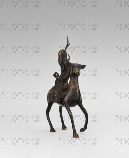 Shoulao on a deer - incense burner, Narozno Eckersdorf, deer, figurines, Chinese art, Chinese culture, incense burner, sages, Taoism, longevity shou iconography, Shouxing iconography, Chinese symbols, Qing 1644–1911, 18th century, China, sculpture, height 27.5 cm, A dark bronze figurine of a person seated on a deer-like animal, with a few areas of worn golden coloring on the surface; the rider has a long flowing beard and elongated headwear with curved horn-like projections, and the cloak and garments drape over the animal's back; the animal has a slender body, long thin legs, small hooves, a narrow face, and upright ears; the object rests on a light neutral background that shifts from pale gray at the top to white at the bottom.