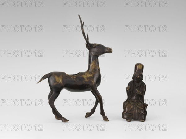 Shoulao on a deer — incense burner; creator, deer and figurines; Chinese art; Chinese culture; Taoist sage Shoulao Shouxing iconography; longevity shou symbol; Qing dynasty 1644–1911, 18th century, China; bronze sculpture; height 27.5 cm, A dark brown metallic deer figure with a few areas of lighter brown patina stands beside a small seated human figure of similar dark brown metal with lighter brown highlights; the deer has an elongated neck, slender legs, a short tail, and tall branching antlers, while the seated figure wears a tall rounded headdress and draped robes and holds its hands near its chest; the background is a pale neutral gradient of light gray and white.