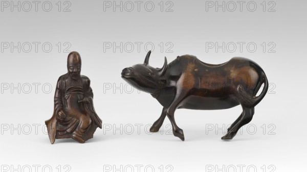 Shoulao on a buffalo - incense burner, Chinese, 19th century Qing 1644–1911, China. Sculpture/figurine depicting Shoulao Shouxing riding a buffalo; Taoist iconography; incense burner. Materials implied sculpture, height 20.7 cm, width 20.7 cm. Subjects: men, buffaloes, sages. Provenance unknown., A small carved figure of a seated person rendered in brown tones with darker brown shading sits to the left, showing a long beard and draped clothing with carved folds; to the right is a carved animal resembling a bovine in polished dark brown and lighter brown areas with curved horns, a rounded body, and slender legs, both objects placed on a white surface against a light gray background.