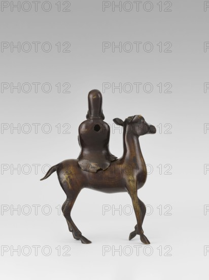 Shoulao on a deer — incense burner; creator, sculptor; Narozno Eckersdorf provenance; deer; Chinese art; Chinese culture; Taoism; incense burner; Shouxing iconography; sages; Qing dynasty 1644–1911, 18th century; China; sculpture; height 23.7 cm, A small sculptural object showing a rider seated on an animal that resembles a camel; the piece appears to be made of a dark brown material with areas of lighter brown and subtle yellowish highlights, and the surface shows a smooth, slightly glossy patina. The rider is simplified and rounded with a hooded or peaked head and a cloak draped over the shoulders, and there is a round opening visible on the rider's back. The animal has a slender neck, an elongated head with small ears, a curved tail, and thin legs with defined hooves, and the overall form shows gentle contours and polished wear on raised areas. The background is neutral and gradient, shifting from pale gray at the top to near white at the bottom.