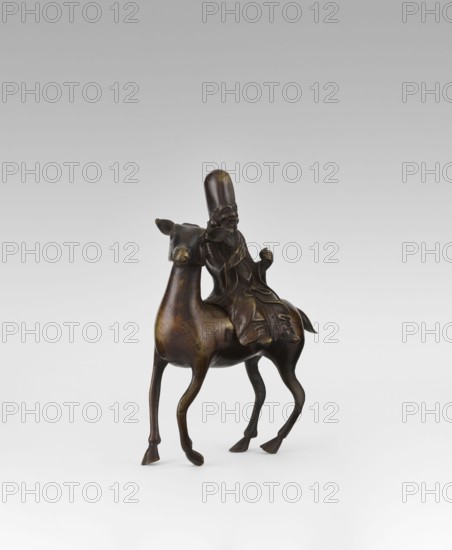Shoulao on a deer — incense burner, deer, Chinese art, Chinese culture, Taoism, incense burner depicting Shouxing and sages, Qing dynasty 1644–1911, 18th century, China, sculpture, height 23.7 cm, A small bronze sculpture shows a robed figure seated on the back of a camel; the figure wears a tall rounded hat and a long flowing robe with carved folds, and the camel has a slender neck, slightly turned head with pointed ears, and long tapered legs. The bronze surface displays dark brown and warm golden-brown tones with subtle polished highlights, and the piece rests on a pale gray to off-white background that gradates to a slightly darker gray toward the top.