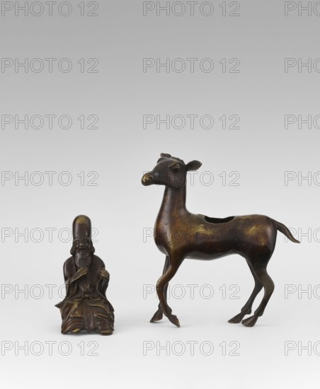 Shoulao on a deer — incense burner, sculptor unknown, 18th century Qing 1644–1911, China; carved/formed sculpture, height 23.7 cm; subject: Shouxing Shoulao riding a deer; Taoist/sage iconography; incense burner; materials implied deer motif, Chinese art., A small dark bronze figure of a seated elderly man with a long beard and tall rounded hat sits beside a larger dark bronze deer with slender legs, a raised head, rounded ears, and a hollowed back; the objects show areas of lighter bronze patina and subtle greenish-gold highlights against an otherwise brown surface, set on a pale gray background.