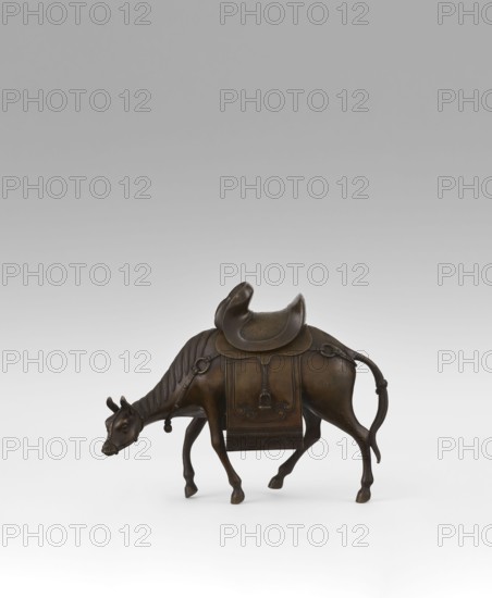 Mule with saddle — incense burner; maker, Qing 1644–1911, 19th century, China; Chinese art; subject: mule with saddle, saddle cloths; material/format: incense burner; height 16.8 cm., A bronze sculpture of a grazing animal with a curved saddle on its back is shown against a neutral gray-to-white background; the animal has a lowered head, slender legs, a long tail with a looped end, and a decorative blanket hanging on its side with ornamental straps and carved details; the surface tones are dark brown with lighter brown highlights and subtle greenish patina in places.