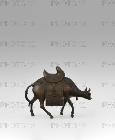 Mule with saddle — incense burner; maker, Qing style; Chinese; subject: mule with saddle, saddle cloths; materials/technique: incense burner; date: 19th century Qing, 1644–1911; height 16.8 cm; provenance unknown., A bronze sculpture of a camel-like animal shown in profile facing right, with a rounded saddle on its back and a decorative blanket hanging down its side; the figure has a curved tail, elongated legs, a lowered head with small curved horns and a ring hanging from its bridle, and the surface displays a dark brown patina against a pale gray to white gradient background.