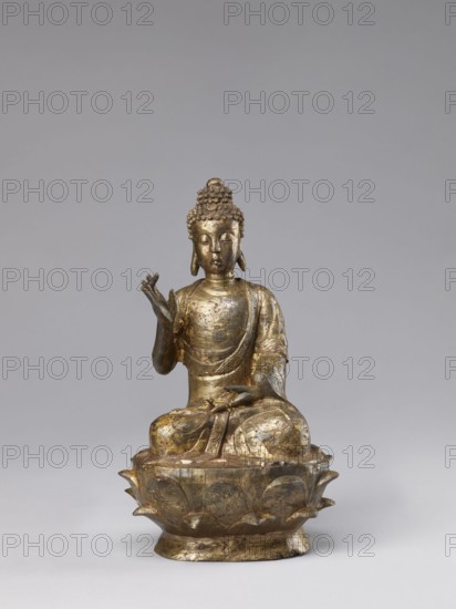 Buddha on a Lotus Throne, Chinese, figurine, Chinese Buddhism, lotus throne iconography, Buddhist art, Qing dynasty 1644–1911, ushnisha, natural patina, 19th century, China, sculpture, height 28.5 cm, A metallic seated figure with a textured hair bun and elongated earlobes sits on a lotus-shaped pedestal, rendered in worn gold and darker tarnished tones; the figure's clothing drapes over the shoulders and knees with visible folds and decorative patterns, showing areas of brighter gold and patches of muted brown and gray; one hand is raised with fingers extended while the other rests on the lap, and the facial expression appears calm with closed eyes; the background is a smooth light gray.