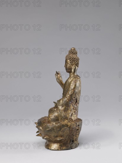 Buddha on a Lotus Throne, Chinese, Chinese art, figurine, Chinese Buddhism, lotus throne iconography, Buddhist art, Qing dynasty 1644–1911, ushnisha, natural patina, 19th century, China, sculpture, height 28.5 cm, A small metallic statue of a seated figure with a textured hair bun and elongated earlobes sits on a rounded base shaped like a stylized lotus, showing worn and corroded surfaces with patches of rough, darkened material and remnants of a golden finish; the figure is shown in profile with one hand raised and the other resting on the lap, wearing draped robes with incised patterns, all set against a smooth, pale gray background.