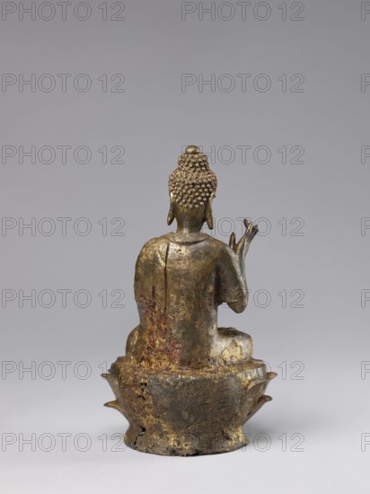 Buddha on a Lotus Throne, Chinese, figurine, Chinese Buddhism, lotus throne iconography, Buddhist art, Qing 1644–1911, ushnisha, natural patina, 19th century, China, sculpture, figurine, height 28.5 cm, A small metallic statue shown from the back, with a textured head of tight curls and a rounded topknot, a pair of elongated ears, and a seated torso draped in a robe. The figure sits on an ornate base with petal-like projections, and the surface shows areas of worn gilding, darkened metal, and patches of reddish and brown discoloration. The background is a smooth neutral gray, and the visible colors include gold, bronze, brown, reddish tones, dark gray, and light gray.