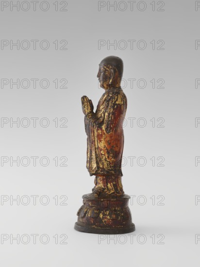 Monk, Chinese, Luohan/arhat figure, monk, sage; Buddhist art; lotus throne iconography; Qing dynasty 1644–1911, 19th century, China; sculpture/figurine; height 24.7 cm., A standing wooden figure shown in profile with hands held together in front, painted in worn shades of gold, dark brown, red, and black, wearing a long robe and standing on a rounded pedestal that shares the same aged gold, red, and dark brown coloration.