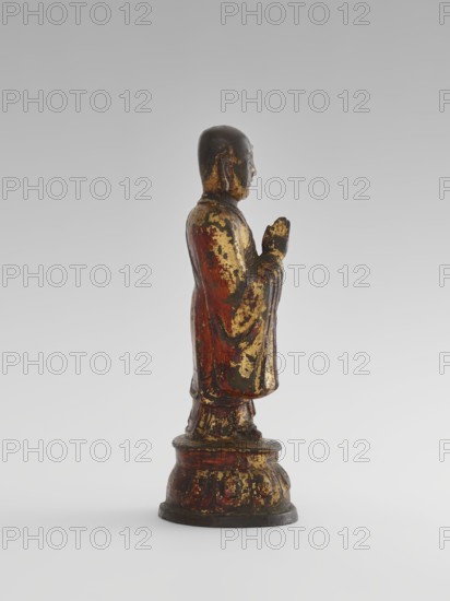Monk, Chinese, Chinese art, sages, Luohan iconography, monks, arhat iconography, Buddhist art, lotus throne iconography, Qing 1644–1911, 19th century, China, sculpture, figurine, height 24.7 cm, A standing figurine shown in profile on a rounded pedestal, with a smooth rounded head and folded hands held near the chest; the surface shows worn patches of gold leaf over a base of reddish-brown and dark brown, with areas of black and muted green where the finish has aged; the robe has flowing vertical folds and the pedestal has carved rounded motifs with similar gold and reddish-brown wear; the background is a smooth gradient of light gray to off-white.
