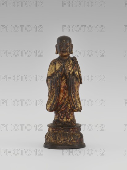 Monk, Chinese, Luohan arhat figure, monk, sage; Buddhist art; lotus throne iconography; Qing dynasty 1644–1911, 19th century; sculpture, figurine; height 24.7 cm, A small standing statue of a robed figure on a rounded base, shown against a neutral gray background; the figure has a rounded head and hands held together near the chest, and the robe and base show worn areas with visible gold and dark brown tones along with traces of red, giving a distressed, aged appearance.
