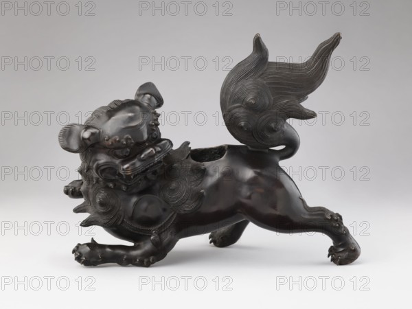 Censer — lion; maker, Chinese; Qing dynasty 1644–1911; 19th century; ceramic/metal? material not specified; ritual/ceremonial vessel; subject: lion/fo iconography; most likely the small lion that forms the cover of the censer is missing; dimensions: height 28.5 cm, width 17.7 cm; origin: China., A dark bronze sculptural figure of an animal with a stylized mane and curled tail is shown in profile on a neutral light gray background, the figure posed with its body low and one front paw extended, the surface smooth and polished with carved swirling patterns on the mane, tail, and shoulder areas, the head featuring prominent rounded ears, a wide open mouth with visible carved teeth, and decorative raised bumps on the face and paws, the overall colors visible are dark brown to nearly black on the sculpture, lighter brown highlights on worn edges, and shades of gray in the background.