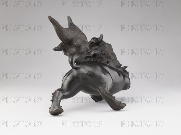 Censer — lion; maker, Chinese; Qing dynasty 1644–1911; 19th century; ceremonial/ritual vessel; subject: lion fo; likely missing small lion that formed the cover; material not specified; dimensions: height 28.5 cm, width 17.7 cm; place of origin: China., A dark, glossy sculptural figure resembling an animal with a rounded body and stylized, flowing mane is shown against a neutral, light gray background. The figure appears to be captured in motion with a raised front limb and a bent rear limb, both ending in rounded feet. The surface of the sculpture is smooth in places and textured with carved, concentric wave-like patterns on the head and mane. Small pointed protrusions decorate parts of the back and limbs. Visible colors in the image include black and dark brown on the sculpture and several shades of gray in the background and surface beneath the object.