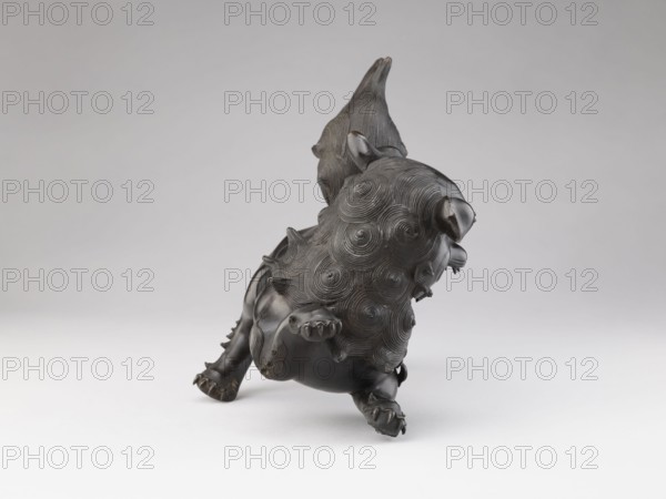 Censer — lion; creator, Chinese; Qing dynasty 1644–1911; ceremonial/ritual vessel; iconography: lion fo; most likely the small lion that forms the cover of the censer is missing; 19th century; China; censer; height 28.5 cm, width 17.7 cm, A dark metallic sculpture of a stylized animal with a large, textured mane composed of concentric swirl patterns, a raised tail with ridged detailing, and pronounced paws with visible claws; the figure rests on a smooth light surface against a pale gray background, and the visible colors are dark bronze or black on the sculpture and shades of light gray and white in the background and surface.