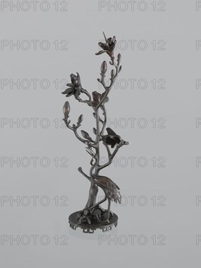 Candlestick shokudai, Japanese, magnolias, Meiji 1868–1912, plant tendril decoration, cranes iconography, 2nd half of the 19th century, Japan, candlestick, height 49.5 cm, The image shows a dark metallic sculpture of a slender tree-like branch with a few blossoms and buds, mounted on a circular base; a bird with a long neck and detailed feathers stands at the base, its body and legs rendered in the same dark metallic material, and the overall colors visible are dark brown and black against a light gray background.