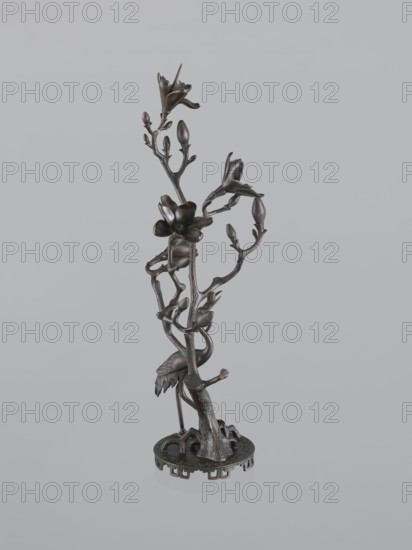 Candlestick shokudai, Japanese, magnolias, Meiji 1868–1912, candlesticks, plant tendril, cranes iconography, 2nd half of the 19th century, Japan, candlestick, height 49.5 cm, A dark metallic sculpture of a slender tree-like stem rising from a round base, with a group of branching twigs and buds and several open blossoms, a bird with a long neck and folded wings standing among the lower branches, and visible textures of bark and feather; the colors visible are dark brown and black against a light gray background.