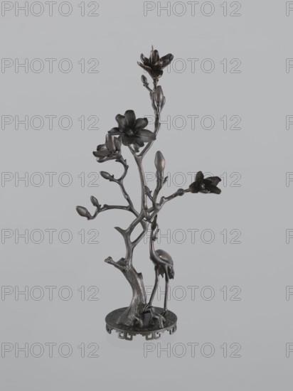 Candlestick shokudai, Japanese, magnolias, Meiji 1868–1912, candlestick with plant tendril and cranes iconography, 2nd half of the 19th century, Japan, height 49.5 cm, A metal sculpture depicting a slender, branching tree with a textured trunk rising from a round base, featuring several stylized blossoms and buds on its branches and a long-legged bird standing beside the trunk; the piece appears in dark bronze tones with hints of metallic sheen against a light gray background.