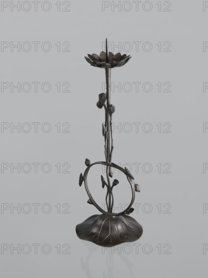 Candlestick shokudai, Takimoto, Iwami; Japanese art; Japanese culture; lotuses, leaves; candlestick; Meiji 1868–1912, 2nd half of the 19th century; Japan; unknown origin; height 44.0 cm, A sculptural metal object resembling a tall, slender stem rising from a rounded, petal-like base with a dark brown to bronze patina, featuring a looping circular vine element mid-height with several small leaf and bud shapes attached, a vertical stem with textured nodes and leaves, and a flower-like dish at the top with radiating petal forms and a pointed central spike; visible colors include dark brown, bronze, and subtle black accents set against a pale gray background.