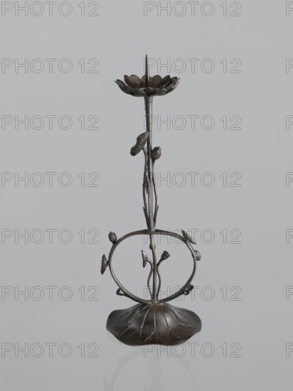 Candlestick shokudai, Takimoto, Iwami; craftsman; Japanese art; Japanese culture; lotuses; leaves; candlesticks; Meiji 1868–1912; unknown origin; 2nd half of the 19th century; Japan; candlestick; height 44.0 cm, A dark metallic decorative stand shaped like a stylized flower and stem sits against a pale gray background; the base resembles a scalloped leaf with visible vein-like ridges, from which a slender textured stem rises and splits into a circular ring adorned with small bud-like and leaf-like elements, and above this the stem continues upward to a bowl-shaped flower top with layered petal forms and a central pointed spike. The visible colors are dark brownish metal tones and shades of gray in the background.