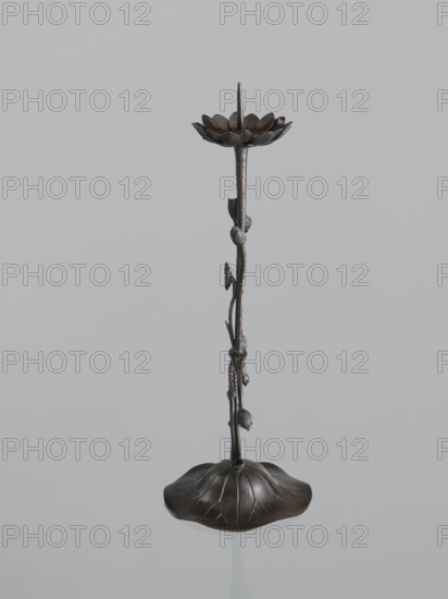 Candlestick shokudai, maker: Takimoto, Iwami; craftsman. Japanese art; Japanese culture. Decorated with lotuses and leaves. Meiji period 1868–1912, 2nd half of the 19th century. Japan. Candlestick, height 44.0 cm. Provenance unknown., A tall, slender metal object shaped like a stem with a flower at the top and a rounded, petal-like base; the surface shows sculpted buds and leaves along the stem and a pointed spike rising from the center of the top flower. The colors visible are dark brown with bronze tones and subtle highlights of lighter brown and black.